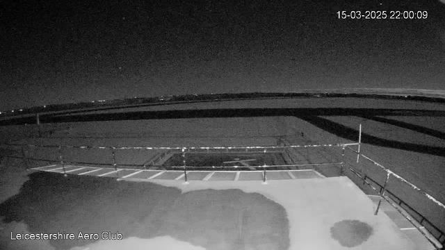 A dark, nighttime scene from a webcam, showing a flat surface with some markings, likely a runway or landing area. In the foreground, there is a railing with horizontal bars, and the background features a flat horizon with a slight incline. The image is monochrome, highlighting the absence of color and the shapes of the surroundings. There is a timestamp indicating the date and time: March 15, 2025, at 10:00 PM.