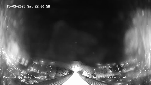 A black and white image captured from a webcam, showing an aerial view of a cityscape at night. The scene is somewhat foggy or hazy, obscuring detailed visibility. Bright lights from buildings and streets are visible in the lower portion of the image, creating a glowing effect on the dark surroundings. The top part features the structure of a roof or ceiling, converging towards a brighter light source in the center, suggesting an opening or sky view. The time and date are displayed in the top left corner. The bottom left corner includes the watermark "Powered By Brighton CCTV."