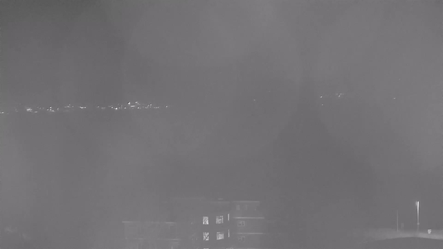 A dimly lit scene showing a cityscape with faint lights in the background, likely from buildings or streetlights. The foreground features a blurred outline of a building. The overall image has a gray tone, giving it a nighttime feel.
