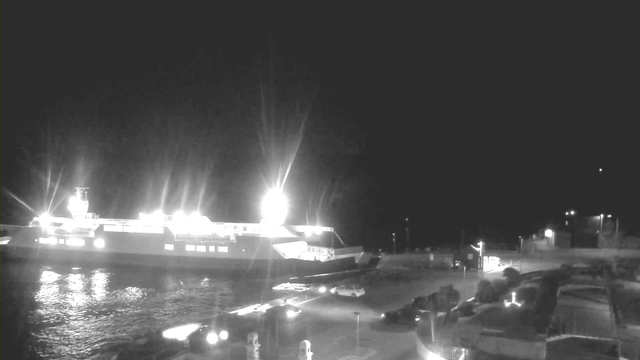 A brightly lit ferry is docked at a harbor during nighttime. The ferry emits strong lights, reflecting off the water. The shoreline features a parking area with some vehicles and dimly lit pathways. There are silhouettes of boats and buildings in the background, creating a tranquil nighttime scene.