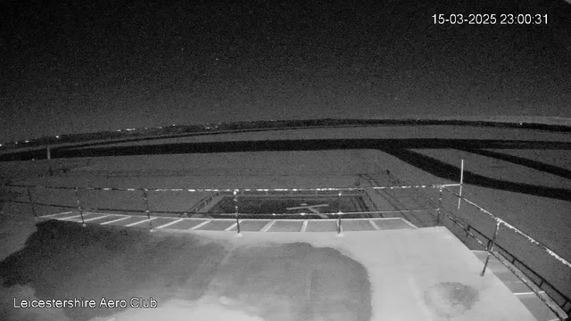 A dimly lit aerial view of an airfield at night. In the foreground, a flat surface with a faint airplane symbol marked in white and a barrier railing along the edge. The background shows an expansive dark sky with a few distant lights, indicating possible buildings or structures far away on the horizon. The atmosphere appears calm and still, with a sense of open space around the airfield.