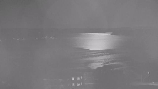 A dimly lit scene of a peaceful body of water under a night sky. The surface of the water reflects light, creating a shimmering effect. In the background, there are faint outlines of hills or land, and a few small lights, possibly from distant buildings or boats, can be seen along the shoreline. In the foreground, shadows suggest structures or vegetation, while a single lamp post is illuminated, casting light onto a nearby area.