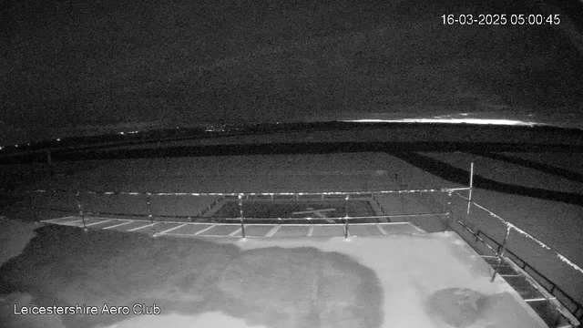 A nighttime image from a webcam at Leicestershire Aero Club, showing a dark sky with some faint lights in the background. The ground is visible with outlines of parking spaces and a helicopter pad marked. The scene is dimly lit, emphasizing the shadows and dark surfaces. The date and time display shows "16-03-2025 05:00:45" in the upper right corner.