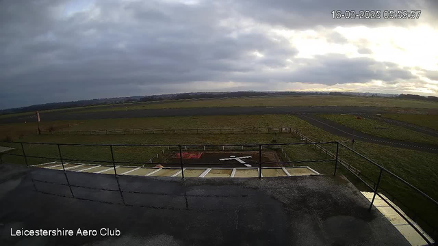 A view from a webcam at Leicestershire Aero Club showing a cloudy sky with varying shades of gray. In the foreground, a railing is visible with a reflective surface. Below, there is a grassy area with a white cross marking, likely a runway or landing area, and some fencing is seen surrounding the perimeter. Beyond the immediate foreground, the field extends outwards towards a distant horizon, with an asphalt runway to the right under the overcast sky. The timestamp indicates the image was taken at 5:59 AM on March 16, 2025.