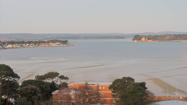 A scenic view of a calm body of water, with gentle ripples and soft reflections. In the foreground, there is a building with a reddish exterior, surrounded by trees. To the left, a shoreline is visible with a few structures and boats. In the distance, hills line the horizon, and a small island with a building sits on the right side of the water, surrounded by greenery. The sky is a light color, suggesting early morning or late afternoon light.