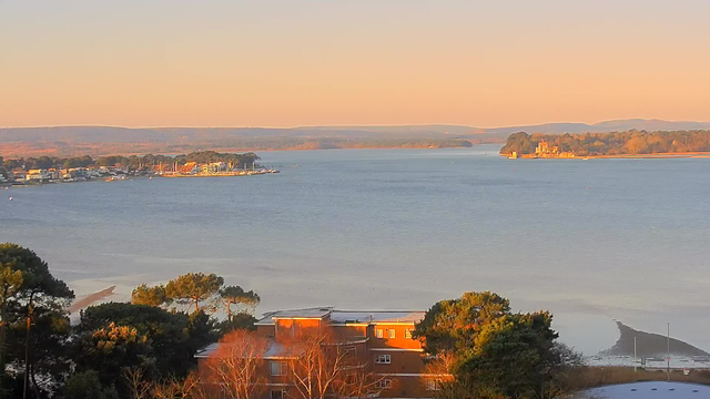A serene view of a body of water reflecting warm colors from a sunset. The shoreline is dotted with trees and buildings, some of which appear to be homes. Small boats are visible near the shore. In the distance, there are hills under a soft, clear sky. The overall scene evokes a peaceful, calm atmosphere.