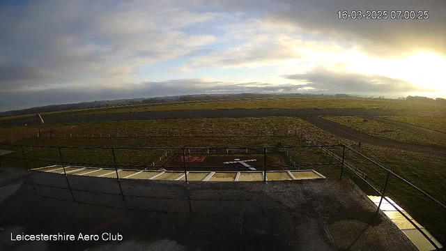 A view from a webcam positioned at an airfield. In the foreground, there is a fenced area with a marked landing cross on the ground. The ground is mostly grassy and slightly damp, visible at the edge of the fence. In the background, there are wide-open fields and a winding asphalt runway. The sky above is cloudy, with patches of sunlight breaking through, hinting at a sunrise. The time stamp indicates it is early morning, and the scene is peaceful with no visible aircraft.