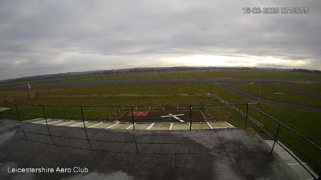 A wide view of a grassy airstrip under a cloudy sky. In the foreground, a wooden railing surrounds a flat surface with a white cross and a red sign on a dark area. The airstrip is lined with grass, and in the distance, there are patches of grey clouds. The scene is relatively empty, conveying a sense of quiet at the Leicestershire Aero Club location.