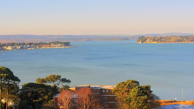 A panoramic view of a calm blue waterscape under a clear sky. In the foreground, there are patches of green trees and a low building. On the left side, a small marina and a village with colorful buildings and boats can be seen along the shore. In the distance, gentle hills rise against the horizon, with more trees lining the water's edge. The light appears warm, suggesting a serene afternoon scene.