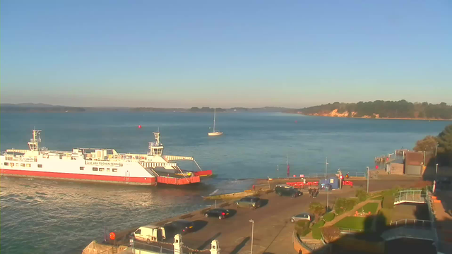 A ferry with a red front is arriving at a pier on a sunny day. The water is calm, and there is a small white sailboat in the background. To the right, there are vehicles parked along the waterfront, and a few people are gathered near the dock. In the distance, a tree-lined shore is visible against a clear blue sky.
