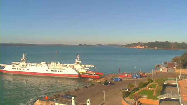 A ferry is docked at a port with a small pier extending into a calm body of water. The ferry is white with red and has a vehicle ramp extended. In the background, there are rolling hills and a clear blue sky. The shoreline features trees and light-colored cliffs. In the foreground, there is a parking area with several cars and a decorative garden with plants. Overall, the scene is bright and sunny.