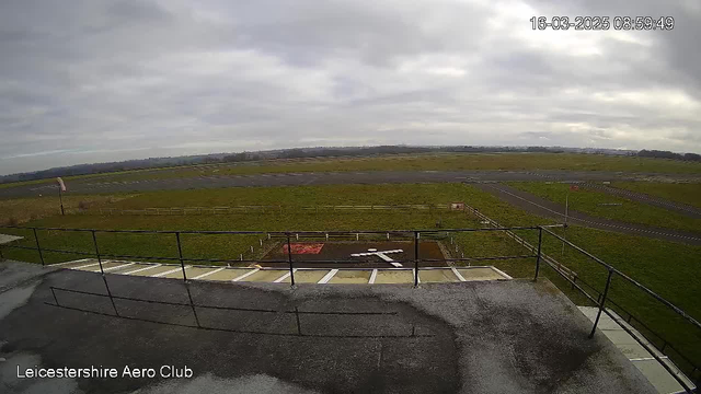 A cloudy sky is visible above an expansive green field with a runway. In the foreground, a balcony railing frames the view. There is a marked helipad with a white cross on a brown surface. Nearby, a sign is partially visible. The scene is set at Leicestershire Aero Club.