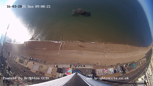 An aerial view of a beach and coastline, with golden sandy shores meeting turquoise waters. In the foreground, there are parts of a building structure with colorful designs. The beach features a pier extending into the water, along with a few people walking along the shore. The water reflects sunlight, creating a shimmering effect. In the distance, remnants of a pier can be seen in the water, while a seaside promenade runs parallel to the beach. The sky is mostly clear, indicating a bright sunny day. The timestamp in the upper left corner reads "16-03-2025 Sun 09:00:21."