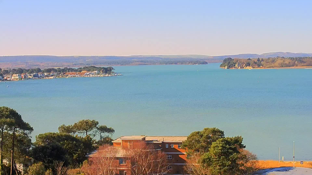 A panoramic view of a tranquil blue body of water, with a shoreline dotted by houses and boats. In the foreground, there are some trees and a brown building. In the background, across the water, there are green hills and scattered vegetation, with a few boats visible on the water. The sky is clear and bright, contributing to a serene atmosphere.