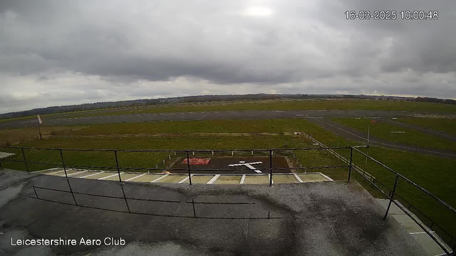 An image from a webcam shows a view of an airfield under a cloudy sky. In the foreground, there is a railing with a flat surface below it, which appears to be a part of a building. The ground in front is grassy, with a red marking and a large white "X" shape, likely indicating a landing area. In the background, the airfield extends with a runway visible and some distant low hills. There are wooden fences bordering the grassy areas, and the overall atmosphere is overcast. The image captures the setting at 10:00 on March 16, 2025.