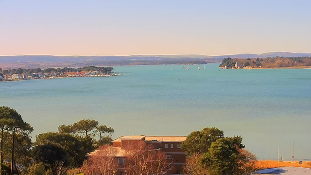 A scenic view of a calm bay with turquoise water. In the foreground, there are trees and a building with a flat roof. Along the left side, small boats are moored in the water, and to the right, several sailboats are visible, sailing across the bay. In the background, hills rise gently, with a few houses scattered along the waterfront. The sky is clear, creating a serene atmosphere.