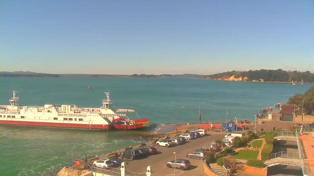 A ferry is docked at a coastal terminal, adjacent to a parking lot filled with cars. The water is a bright turquoise, with gentle waves and a few small boats visible further out. The background features a shoreline with trees and a small hill. The sky is clear and blue, indicating a sunny day. The scene is peaceful and captures the essence of a seaside location.