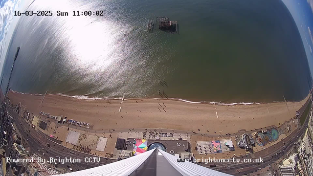 A high-angle view of a beach with golden sand and a wide stretch of blue ocean. The shoreline is lined with light waves and there are several people walking along the beach. In the foreground, there are colorful beach amenities, including a circular pool and a multi-colored geometric pattern on the ground. To the left, remnants of a pier can be seen extending into the water. The sky above is partly cloudy with bright sunlight reflecting off the ocean surface. The date and time display in the top corner shows "16-03-2025 Sun 11:00:02."