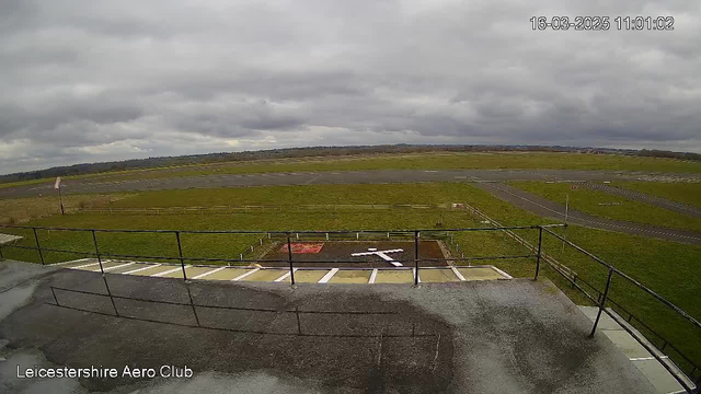 A view from a webcam at Leicestershire Aero Club showing an expansive grassy area and a small runway under a cloudy sky. In the foreground, there is a railing and some concrete steps leading down from a building. On the ground, a white cross shape and a red shape are visible in the grass, indicating a designated landing area. The background features more grassy fields and a distant horizon, with limited visibility due to the overcast sky.