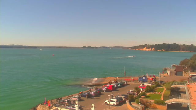 A scenic view of a coastal area on a clear day. The foreground features a parking lot with several parked cars in various colors. A pathway leads down to a small beach area with gentle waves lapping at the shore. In the background, the turquoise water extends towards the horizon, where there are distant hills and a shoreline dotted with trees. A small boat is visible on the water, with other boats in the distance. The sky is bright blue with a few scattered clouds.