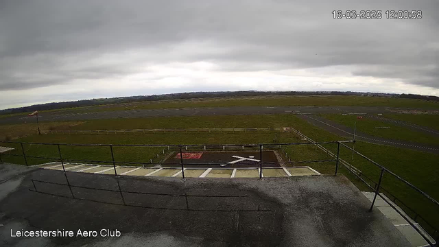 A view from a high vantage point overlooking a grassy airfield under a gray sky. The foreground features a railing and a concrete surface, with a red and white marked area on the ground that includes a large white X. In the distance, a runway lined with fences shows a patch of grass and a partially cloudy horizon. The image is timestamped with the date and time.