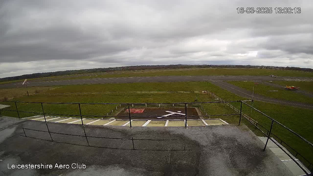 A webcam view from a high vantage point overlooking a grassy area with an airstrip. The foreground includes a railing and steps leading down. In the background, there are clouds in a gray sky, with patches of blue. The airstrip is visible, bordered by fences, and a small aircraft is parked to the right. A red area on the ground has a white cross marked on it. The scene appears overcast and features a sprawling landscape beyond the airstrip.