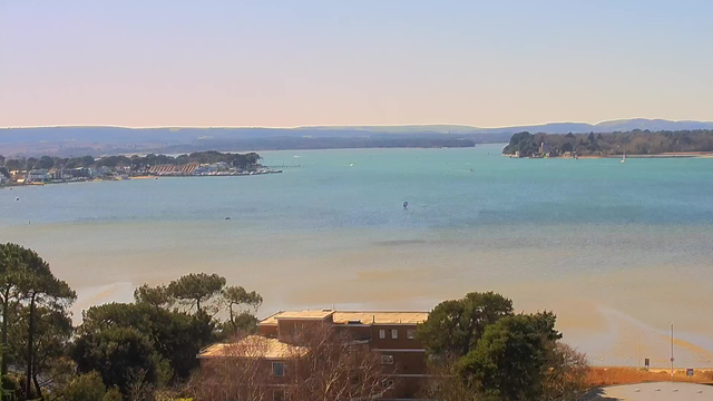 A scenic view of a calm bay with tranquil blue and green waters. In the foreground, there are some trees and a brown building, partially obscured by foliage. To the left, a marina can be seen with small boats docked. The opposite shore features a sandy beach area with a few structures. In the background, low hills create a gentle horizon under a clear sky.