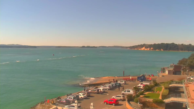 A coastal scene featuring a bright blue sea under a clear sky. In the foreground, there is a parking lot filled with various cars, including white and red vehicles. A walkway leads down toward the water where people can be seen. In the distance, gentle hills and a sandy shore are visible, with some greenery along the coastline. The water shows light waves and traces from boats.