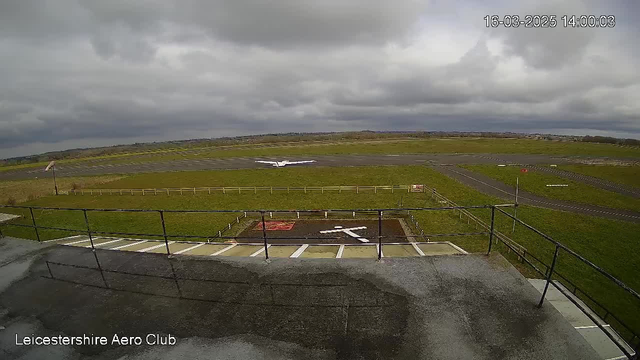 A view from a webcam located at Leicestershire Aero Club. The foreground shows a balcony railing and some ground surface; in the background, a small airplane is taking off on a grass airstrip. The sky is overcast with gray clouds, and a few green fields are visible surrounding the airstrip. There are runway markings and a windsock indicating wind direction.