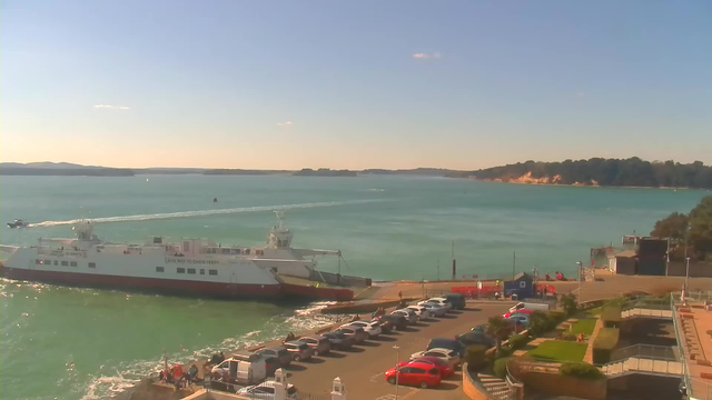 A serene waterfront scene captures a ferry docked at a harbor under a clear blue sky. The water reflects shades of turquoise, while a smaller boat creates a wake as it moves away. In the foreground, there is a long line of parked cars along the harbor, with shrubs and steps leading to a more developed area with a building. The distant shoreline features trees and sandy cliffs, completing the peaceful coastal landscape.