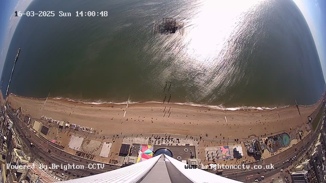 Aerial view of a sandy beach in Brighton, with people walking along the shore. The sea is visible, reflecting sunlight, with waves gently lapping the sand. Large amusement rides are located near the beach, and colorful beach huts line the promenade. A pier extends into the water on the left side of the image. The date and time are displayed at the top of the image.