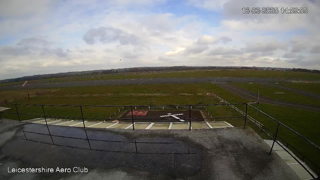 A view from a webcam at Leicestershire Aero Club, showing an expansive grass airfield under a cloudy sky. In the foreground, there is a railing with a gray surface beneath it. To the right, there is a faint white marking on the ground, likely a runway symbol, with a red section beside it. The background features a long runway stretching towards the horizon, bordered by a grassy area and some distant hills. The overall atmosphere appears calm and overcast. The time displayed is 14:59:55 on March 16, 2025.