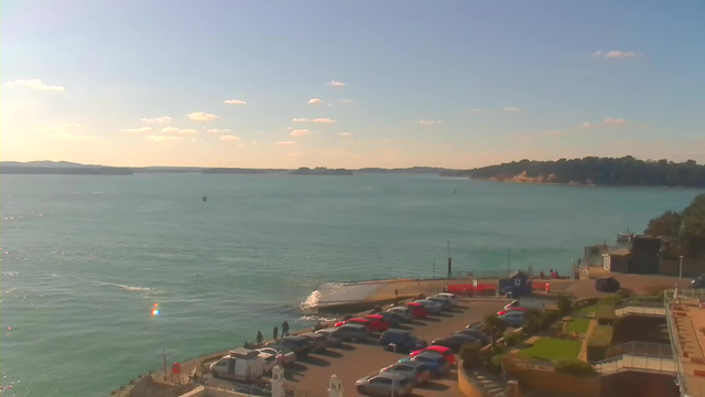A scenic waterfront view featuring calm turquoise waters under a bright blue sky with a few scattered clouds. In the foreground, there's a parking lot filled with various cars, and a few people walking near the edge of a pier. The background showcases distant hills and a rocky shoreline. The sunlight reflects off the water, creating a shimmering effect.