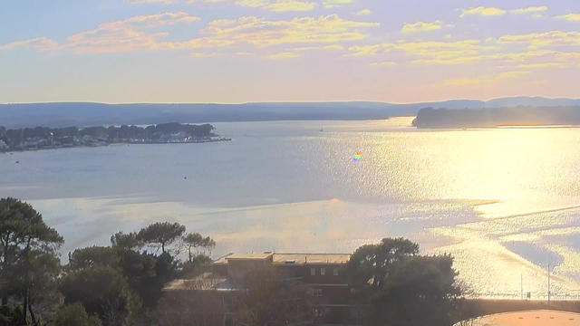 A serene view of a calm body of water, reflecting soft, golden sunlight. In the distance, trees line the shore, and a small town can be seen along the water's edge. Gentle ripples disturb the surface, while a few boats float quietly. The background features rolling hills under a clear sky, with scattered clouds adding to the tranquil atmosphere.