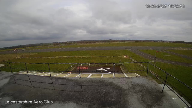 A view from a webcam at an airfield, showing a large, grassy area with a runway. In the foreground, there is a flat roof with railing. On the ground, there is a marked helipad with a white cross. The sky is overcast with gray clouds, and there is a distant horizon line with more fields and a few structures in the background. The image timestamp indicates it was taken in the afternoon.