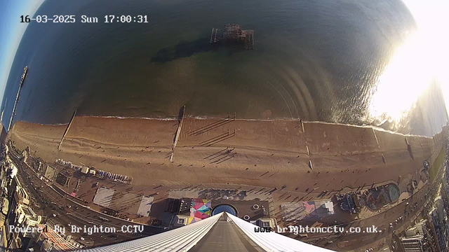 Aerial view of Brighton beach taken from a webcam. The image shows a sandy beach with scattered shadows and people walking along the water's edge. In the distance, there is an abandoned pier jutting into the sea, while gentle waves lapping at the shore create ripples in the water. The sun reflects brightly off the ocean surface, making it difficult to see in that area. Below the beach, a colorful amusement area is visible, including patterns on the ground and structures hinting at rides or activities. The date and time are displayed in the upper left corner.