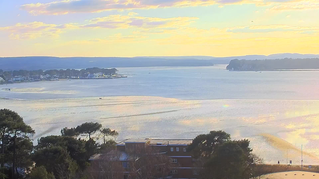 A tranquil view of a wide water body stretching into the distance under a soft, pastel sky. The horizon features gentle hills with some trees and buildings along the shoreline. Reflections of the sky can be seen on the water's surface, which has small ripples. A few boats can be spotted on the water, and the scene conveys a peaceful, serene atmosphere.