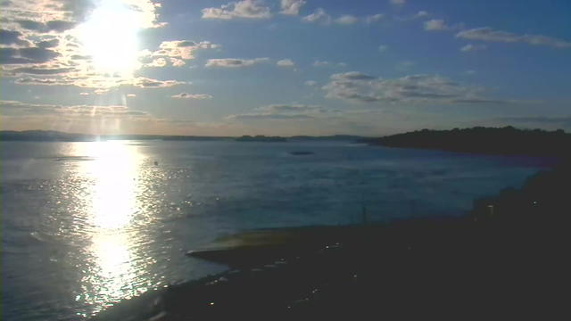 A serene waterfront scene under a partly cloudy sky. The sun is low on the horizon, casting a bright reflection on the calm water, creating a shimmering effect. In the distance, the outline of a landmass can be seen, with a few small boats on the water. The foreground features a dark shoreline, suggesting rocks or a pier, adding contrast to the bright water and sky.