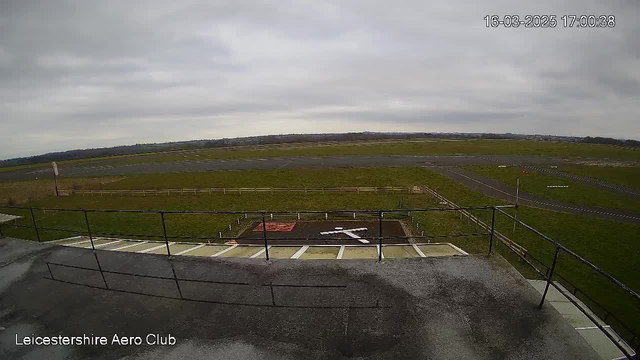 A wide view of an airport runway captured from a webcam. In the foreground, there is a flat roof with a railing. Below, a grassy area extends toward a tarmac runway, which is visible in the background. The sky is overcast with gray clouds, and a faint outline of distant hills can be seen on the horizon. A white cross is painted on the ground, indicating a landing area. The image is timestamped at 17:00 on March 16, 2025.