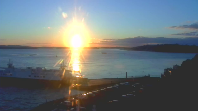 A sunset over a calm body of water. The sun is bright and large, casting a golden reflection on the surface of the water. Silhouetted against the colorful sky, there are dark hills in the distance. In the foreground, a ferry boat is docked near a parking area with several cars visible. The colors in the sky transition from golden yellow near the sun to soft blue and hints of orange as the sky stretches. The scene evokes a tranquil and scenic evening atmosphere.