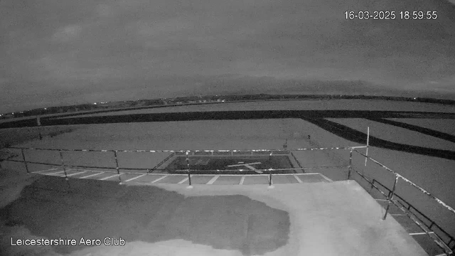 A dimly lit image shows a helipad with an outlined cross pattern in the center, situated on a flat surface. Surrounding the helipad is a grid of white lines, indicating parking spaces or pathways. In the background, there is a dark sky with clouds, and faint lights from distant buildings or structures can be seen beyond the helipad. The time and date are displayed in the top right corner, reading 18:59:55 on March 16, 2025. The bottom left corner includes text identifying the location as "Leicestershire Aero Club."