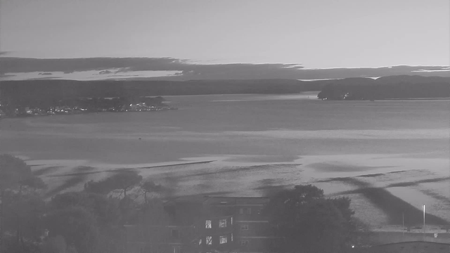 A black and white image of a calm body of water stretching out to the horizon. In the background, there are distant hills partially covered by clouds. The shoreline is visible on the left, with some trees and buildings in the foreground. The water reflects light softly, with subtle ripples creating a serene atmosphere.