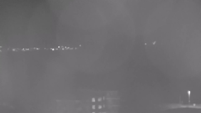 A night scene captured by a webcam, featuring a dimly lit urban landscape. Blurry lights from distant buildings and street lamps create a sparse, sparkling effect against a dark background. The overall image is grainy and lacks detail, suggesting a foggy or low-visibility condition.