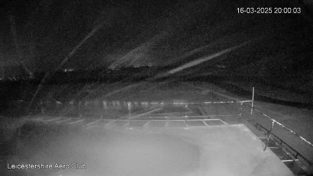 A dark, unclear image from a webcam showing a night scene at Leicestershire Aero Club. There is a parking area with some faint lighting visible in the distance. The sky appears cloudy with streaks of light suggesting either weather activity or reflections. The timestamp indicates the date and time as March 16, 2025, at 8:00 PM.