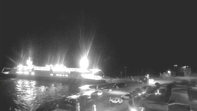 A nighttime image shows a large ferry docked at a waterfront. The ferry is illuminated with bright lights, creating reflections on the calm water. In the foreground, there is a parking area with several vehicles, including white and dark cars. The background features a dimly lit shoreline and a few scattered lights, hinting at structures or buildings nearby. The overall scene conveys a serene nighttime atmosphere at a port.
