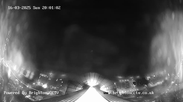 A black and white webcam image showing a cityscape at night. The view is somewhat hazy with bright lights visible in the lower part of the image, indicating urban surroundings. There is a central bright area that suggests a light source or reflection. The date and time are displayed in the top left corner, showing it is March 16, 2025, at 20:01:02. The bottom left corner includes the text "Powered By Brighton CCTV" and the website URL "www.brightoncctv.co.uk."
