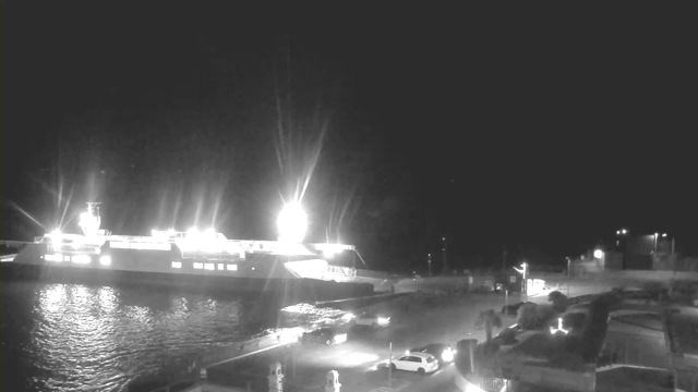 A brightly lit ferry is docked at night, with bright lights illuminating its upper deck. The water reflects the light, creating a shimmering effect. In the foreground, several vehicles are parked and appear to be moving near the waterfront. The surrounding area has some buildings and streetlights, suggesting a port environment at night.