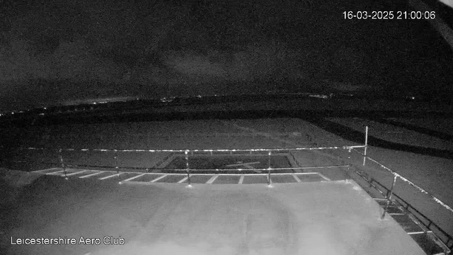 A dimly lit view from a webcam at the Leicestershire Aero Club shows a flat surface with faint outlines of a runway. A railing partially obstructs the view, and dark clouds obscure any light sources in the night sky. The time stamp indicates it is 21:00 on March 16, 2025. The landscape beyond the runway is mostly dark with a few distant lights visible.