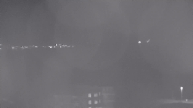 A dark image depicting a nighttime scene with faint lights in the background. Vague outlines of buildings are seen at the bottom, and scattered small, bright dots that may indicate distant streetlights or reflections are present in the upper part of the image. The overall tone is monochromatic, suggesting low visibility.