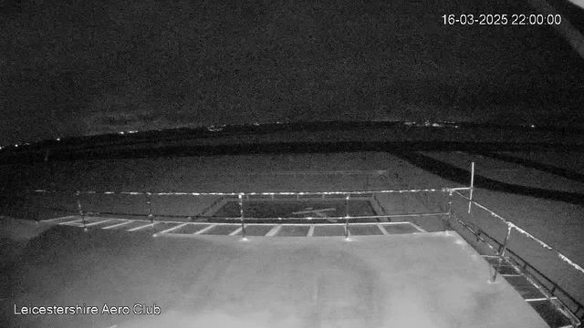A dark webcam image showing a view from the Leicestershire Aero Club. The foreground features a flat surface, possibly a runway or tarmac, with a fenced area along one side. In the distance, faint lights indicate a horizon against a night sky, with clouds partially obscuring visibility. The time and date are displayed in the top right corner, showing 22:00 on March 16, 2025.